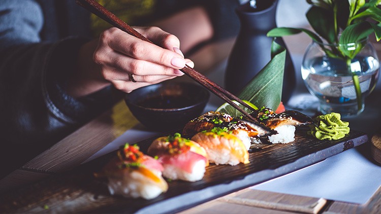Eine Hand hält Essstäbchen und nimmt ein Sushi-Stück von einer schwarzen Platte mit Sushi, Wasabi und grüner Dekoration.