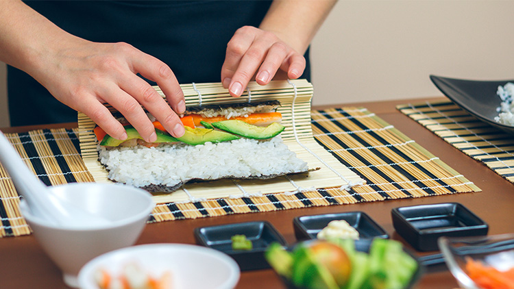 Hände rollen eine Sushirolle aus Seetang, Reis, Avocado und Karotten auf einer Bambusmatte.
