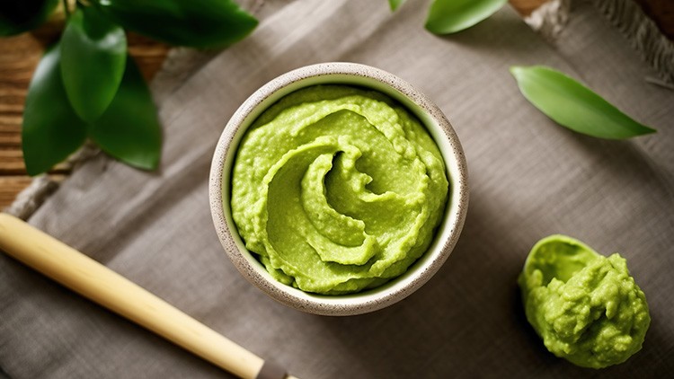 Eine Schale mit grüner Avocadocreme steht auf einem beigen Tuch, daneben liegt ein Messer mit Holzgriff und etwas Creme.