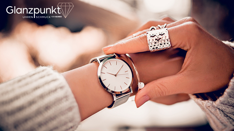 Eine silberne Armbanduhr mit weißem Zifferblatt wird an ein Handgelenk gelegt, oben rechts steht „Glanzpunkt Uhren Schmuck von GLOBUS“.