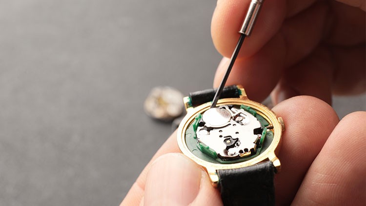 Eine Person öffnet mit einem feinen Werkzeug eine Armbanduhr, das Uhrwerk und die Batterie sind sichtbar.