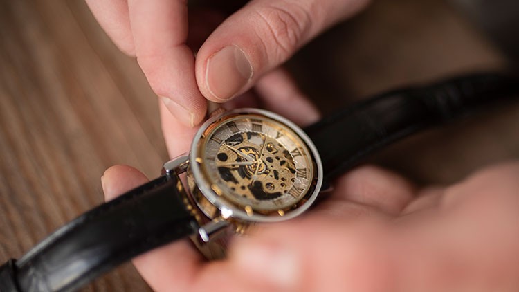 Eine goldene Armbanduhr mit schwarzem Lederband wird von zwei Händen über einer Holzoberfläche gehalten.