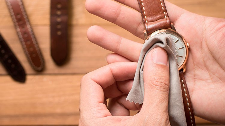 Eine Person reinigt eine Armbanduhr mit braunem Lederarmband und weißem Zifferblatt mittels eines grauen Tuchs.