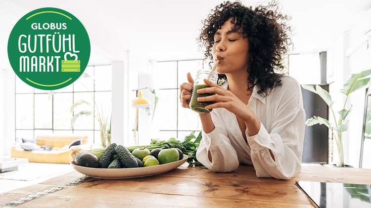 Person in weißem Hemd hält ein Glas mit grünem Smoothie, links oben das grüne Logo „GLOBUS GUTFÜHL MARKT“.