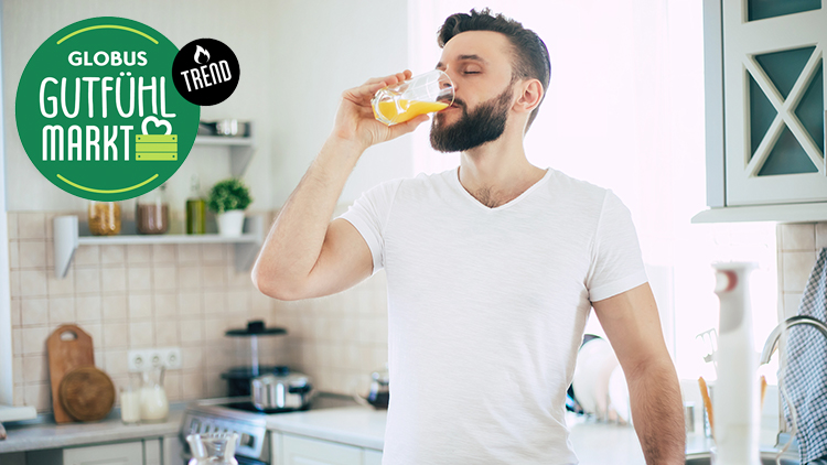 Person in weißem T-Shirt trinkt aus einem Glas mit orangener Flüssigkeit, im Hintergrund eine Küche mit Regal und Schränken.