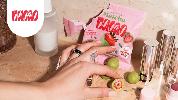 Hand mit Tattoo greift nach grünem Snack neben rosa Verpackung mit rotem Schriftzug „nucao“ und Matcha-Design.