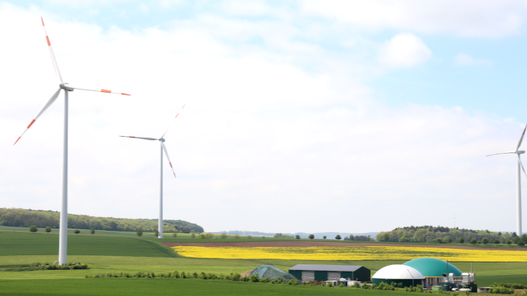 Drei Windkraftanlagen stehen auf einer grünen Landschaft mit Feldern, Bäumen und einer Biogasanlage im Vordergrund.