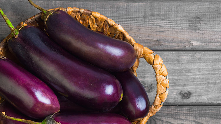 Mehrere violette Auberginen liegen in einem geflochtenen Korb auf einer grauen Holzfläche.