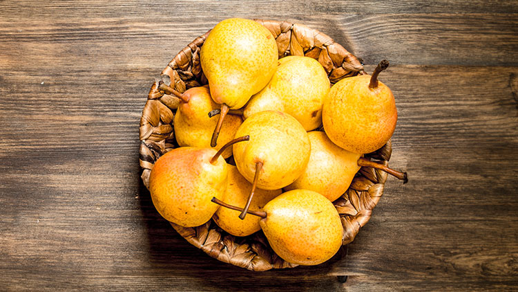 Gelbe und orangene Birnen liegen in einem geflochtenen Korb auf einem Holztisch mit sichtbarer Holzmaserung.