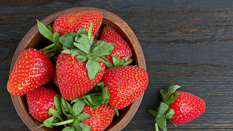 Rote Erdbeeren mit grünem Blattansatz liegen in einer braunen Holzschale auf einer dunklen Holzoberfläche.