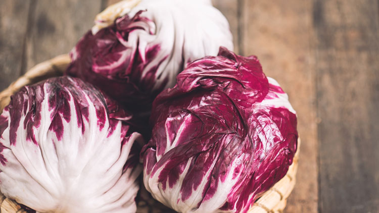 Drei Köpfe Radicchio liegen in einem geflochtenen Korb auf einer rustikalen Holzoberfläche.