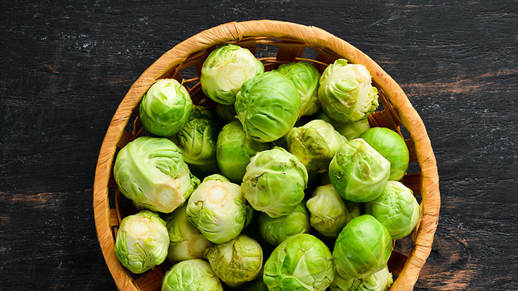 Ein geflochtener Korb mit frischem, grünem Rosenkohl, liegend auf einer dunkel gemaserten Holzoberfläche.