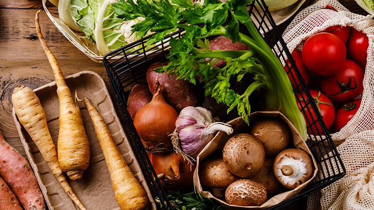 Frisches Gemüse wie Pilze, Süßkartoffeln, Sellerie, Tomaten, Zwiebeln, Pastinaken und Knoblauch liegen in Körben auf Holzuntergrund.