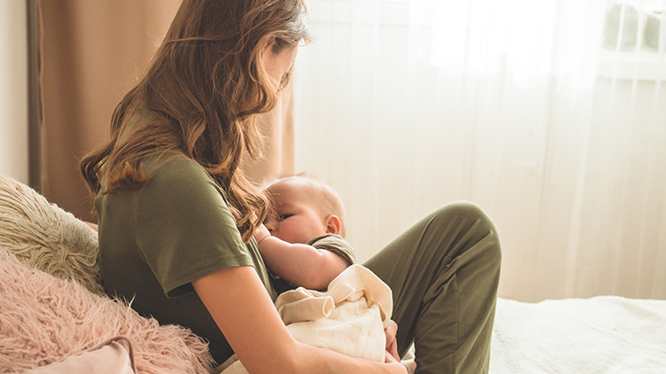 Eine Person in grüner Kleidung sitzt auf einem Sofa und hält ein Baby in den Armen, das an ihrer Brust trinkt.
