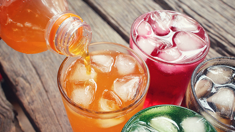 Ein Glas mit orangefarbenem Getränk wird eingeschenkt, daneben stehen drei Gläser mit roten, grünen und klaren Getränken und Eiswürfeln.