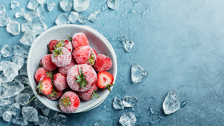 Gefrorene Erdbeeren mit Frostschicht liegen in einer weißen Schale, umgeben von lose verteilten Eiswürfeln auf blauem Untergrund.