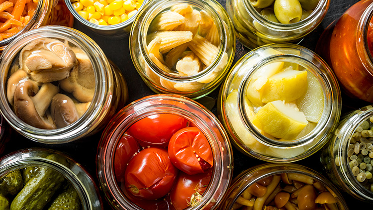 Verschiedene glasgefüllte Lebensmittel wie Mais, Pilze, Oliven, Babymais und Paprika in Nahaufnahme von oben fotografiert.
