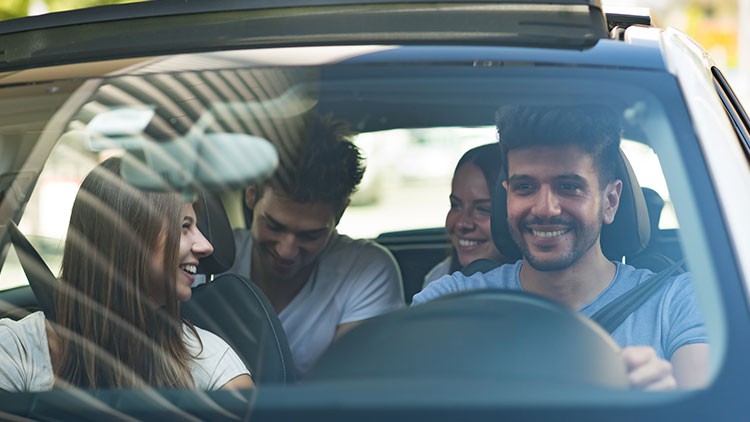 Drei Personen sitzen in einem Auto, zwei auf den Vordersitzen und eine auf dem Rücksitz, bei offenem Schiebedach.