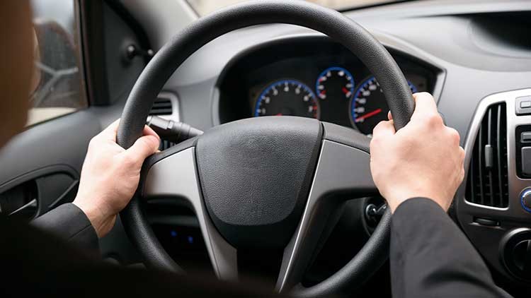 Hände halten ein schwarzes Lenkrad, im Hintergrund sind ein Tachometer, weitere Anzeigen und Fahrzeugbedienelemente sichtbar.