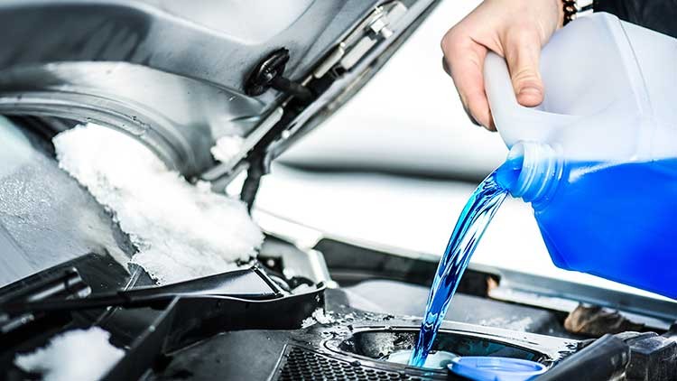 Eine Hand gießt blaue Flüssigkeit aus einem Kanister in den Behälter einer Motorhaube, auf der Schnee liegt.