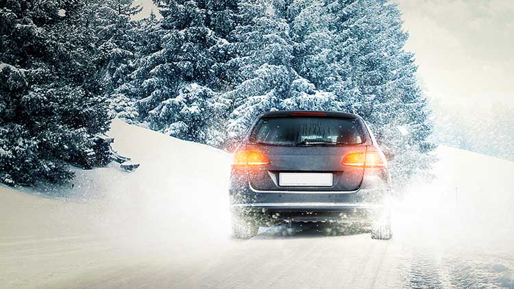 Ein dunkles Auto fährt auf einer verschneiten Straße durch eine winterliche Landschaft mit schneebedeckten Bäumen.