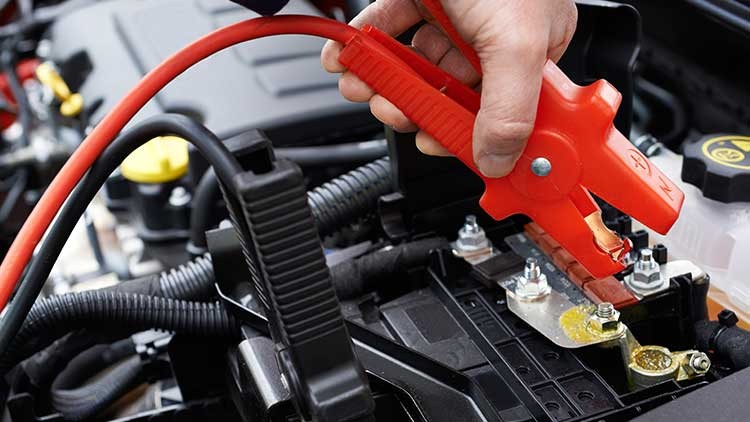 Eine Hand hält ein rotes Überbrückungskabel, das an den Pluspol einer Autobatterie angeschlossen wird.
