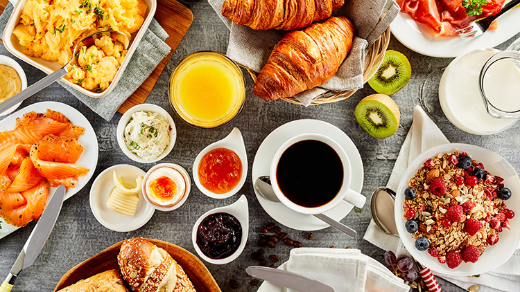 Ein Frühstück mit Rührei, Croissants, Lachs, Obst, Müsli, Kaffee, Orangensaft, Brotaufstrichen sowie Milch und Kiwi.