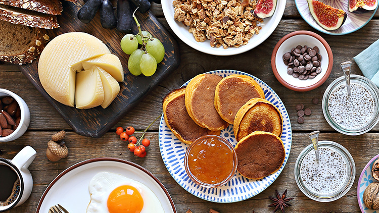 Verschiedene Frühstückszutaten wie Pancakes, Chiasamen-Pudding, Marmelade, Käse, Walnüsse und ein Spiegelei auf Holztisch.