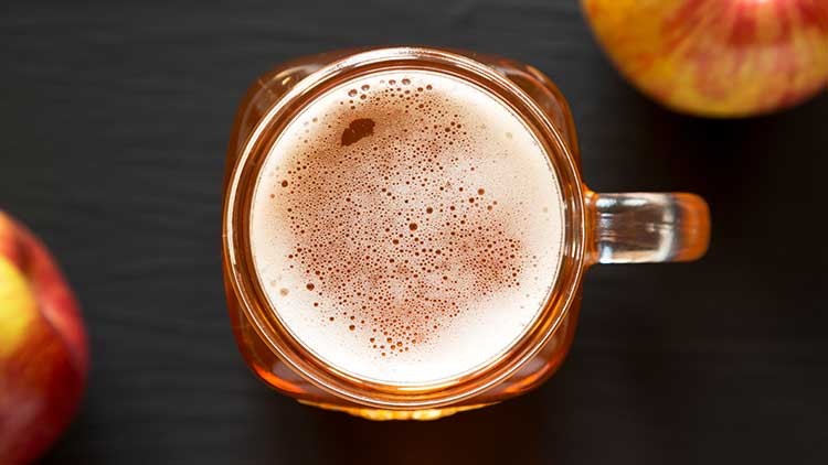Ein Glas mit Schaum bedecktem, hellbraunem Getränk steht auf dunklem Untergrund, daneben zwei rot-gelbe Äpfel.
