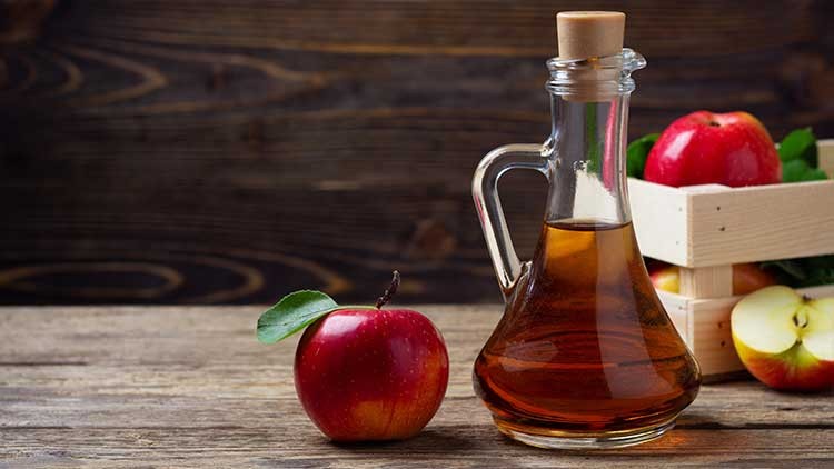 Eine Glaskaraffe mit Apfelessig steht neben einem roten Apfel und einer Holzkiste mit weiteren Äpfeln auf einem Holztisch.