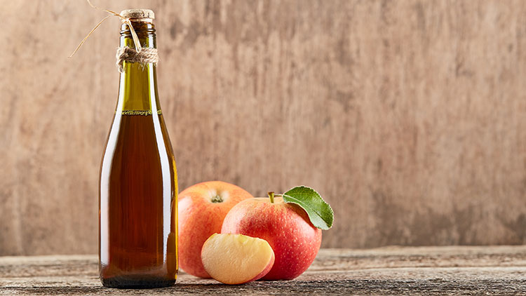 Braune Flasche mit Apfelsaft steht auf Holzuntergrund, daneben zwei rote Äpfel und ein halber angeschnittener Apfel.