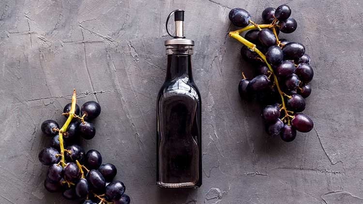 Eine dunkle Glasflasche mit Metallverschluss steht zwischen zwei Bündeln dunkler Weintrauben auf grauem Untergrund.