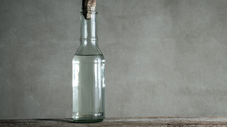 Eine durchsichtige Glasflasche mit klarem Inhalt steht auf einer hölzernen Oberfläche vor einer grauen Wand.
