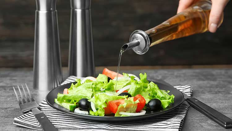 Eine Hand gießt dunkles Dressing aus einer Flasche über einen frischen Salat mit grünem Blattgemüse, Tomaten und Oliven.
