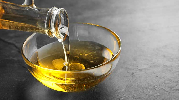 Goldgelbes Öl wird aus einer gläsernen Flasche in eine kleine, transparente Glasschale auf dunklem Untergrund gegossen.