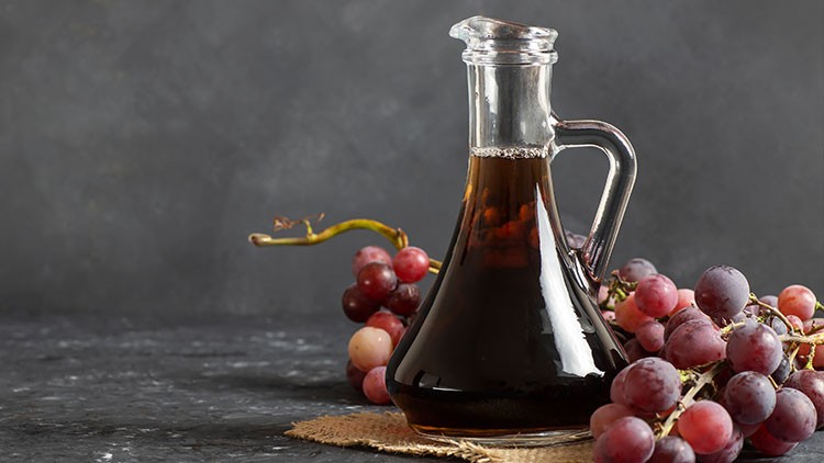 Glaskaraffe mit dunklem Essig auf einem runden Jutestück, dekoriert mit roten und violetten Weintrauben vor dunklem Hintergrund.