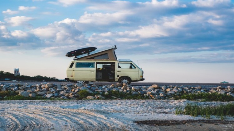Ein beigefarbener Campingbus mit aufgeklapptem Dach parkt auf einem Schotterweg vor einem bewölkten Himmel.