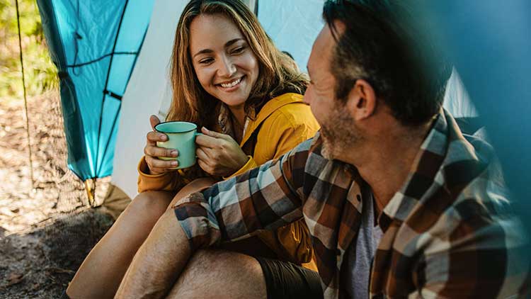 Person in gelber Jacke hält eine grüne Tasse vor einem blauen Zelt, im Hintergrund sind Bäume und unscharfes Laub zu sehen.