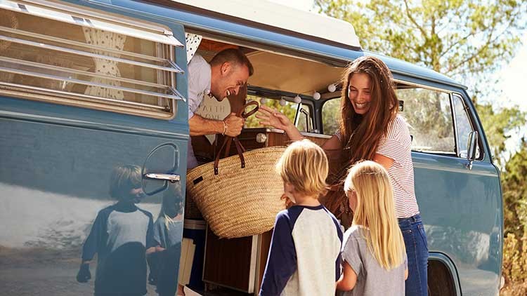 Ein blaues Campingfahrzeug steht offen, während Personen eine Tasche einladen, zwei Kinder stehen davor und blicken hinein.