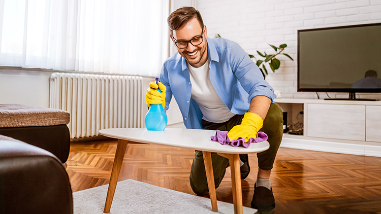 Person mit gelben Handschuhen reinigt einen weißen Tisch mit lila Tuch und blauer Sprühflasche in einem Wohnraum.