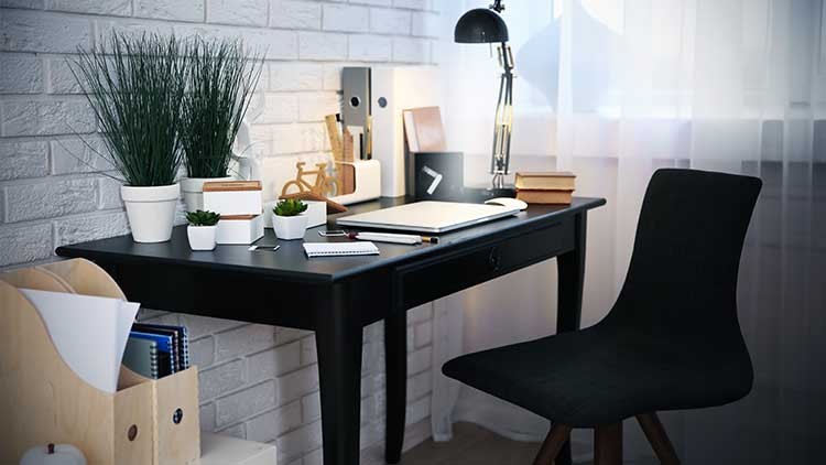 Schwarzer Schreibtisch mit Lampe, Büchern, Pflanzen in weißen Töpfen und Schreibmaterialien vor einer weißen Ziegelwand.