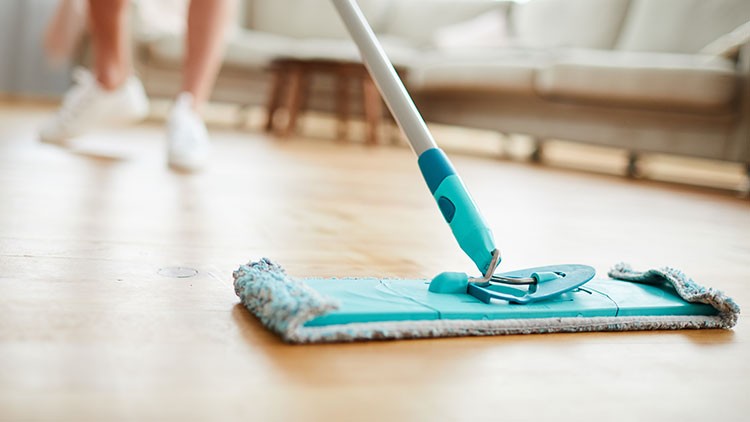 Eine Person wischt mit einem blauen Wischmopp einen hellen Holzfußboden, im Hintergrund ist ein Sofa unscharf erkennbar.