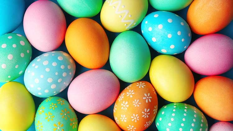 Bunte Ostereier in verschiedenen Farben mit Mustern wie Punkten, Streifen und Blumen liegen eng beieinander auf einer Fläche.