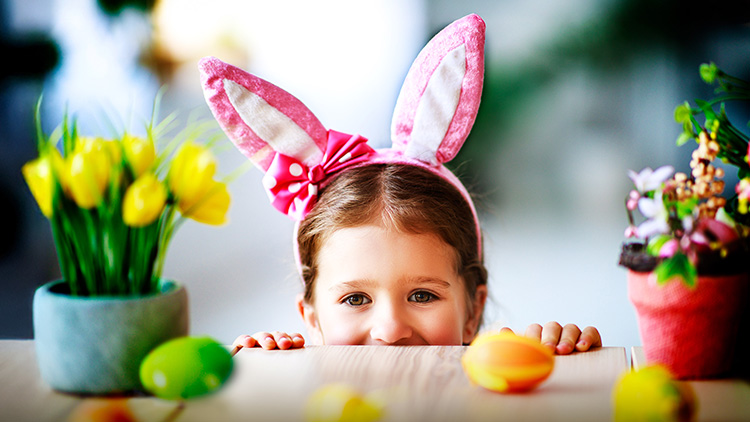 Zwischen gelben und bunten Frühlingsblumen liegen farbige Ostereier auf einem Tisch, im Hintergrund verschwommene Dekoration.