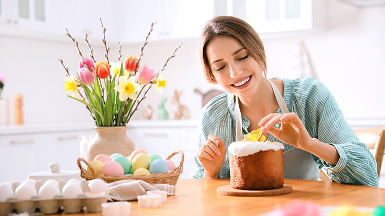 Person dekoriert einen Kuchen mit Glasur, bunte Eier in einem Korb, Tulpen und Weidenkätzchen in einer Vase auf einem Holztisch.