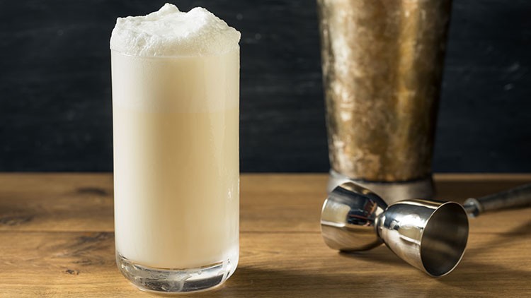 Ein hohes Glas mit weißem, schaumigem Getränk steht auf einem Holztisch, daneben ein Edelstahldosierer und ein Shaker.
