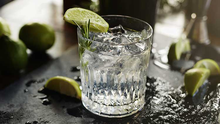 Ein Glas mit Eiswürfeln, Limettenscheibe und Rosmarinzweig, umgeben von frischen Limetten und tropfnassen Untergrund.