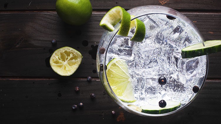 Ein Glas mit Eiswürfeln, Gurkenscheiben, Limettenspalten und Wacholderbeeren auf dunklem Holzuntergrund, daneben halbe Limette.