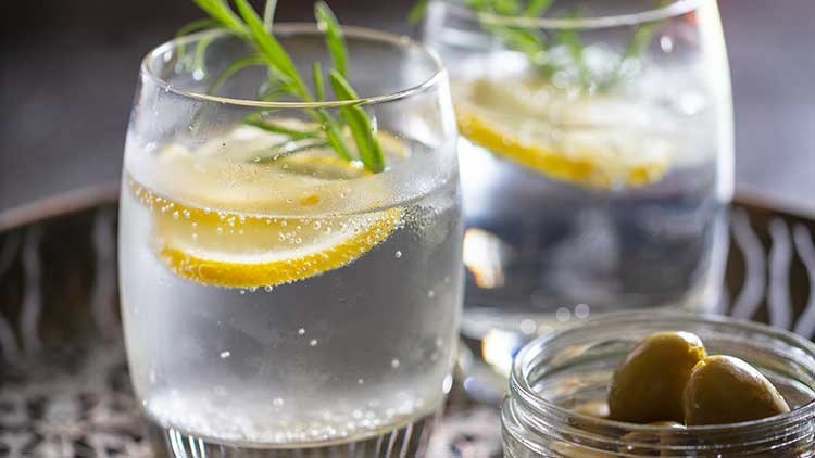 Zwei Gläser mit durchsichtigem Getränk, Zitronenscheiben und Rosmarinzweigen, daneben ein Glas mit grünen Oliven.