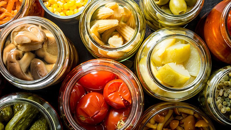 Verschiedenes eingelegtes Gemüse wie Pilze, Tomaten, Gurken, Mais, Babymais, Oliven und Paprika in Gläsern von oben gesehen.